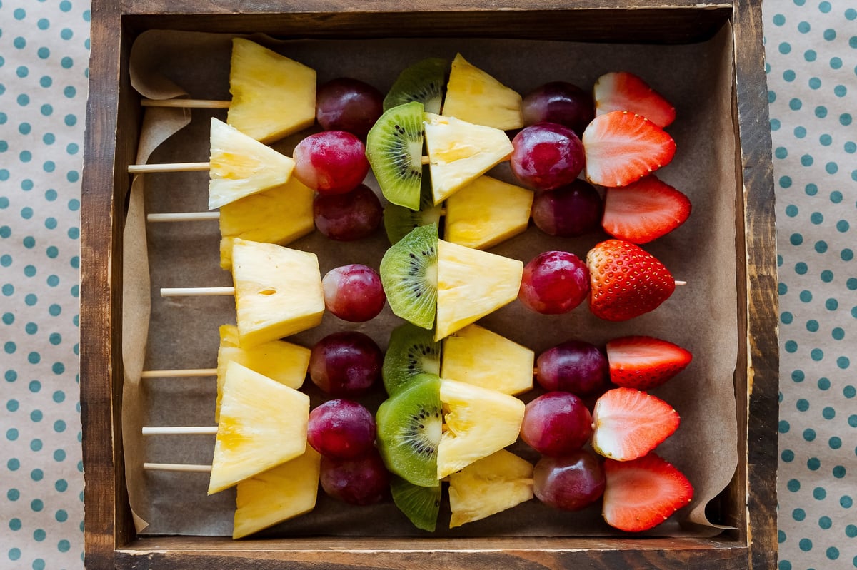 A crafted wooden box contains colorful skewers filled with fresh fruit like pineapple, juicy grapes, vibrant kiwi, and sweet strawberries